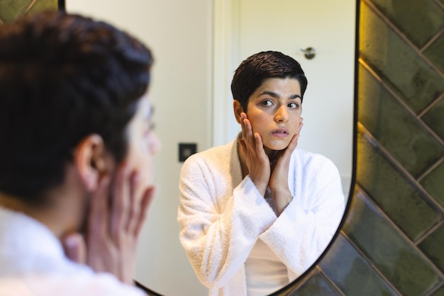 Happy biracial woman touching face looking at reflection in bathroom mirror at home