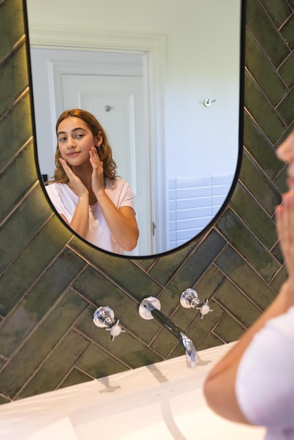 Happy biracial woman touching face and smiling looking at reflection in bathroom mirror at home