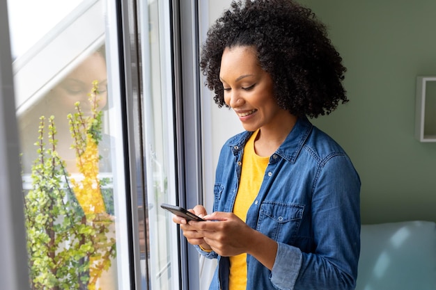 happy biracial woman using smartphone by window at home. lifestyle, communication and domestic life, unaltered.