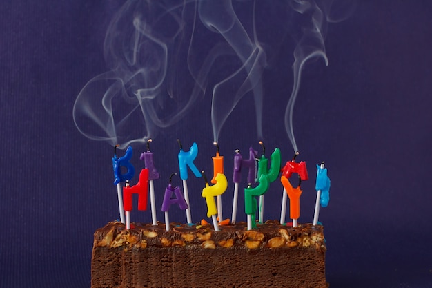 お誕生日おめでとうブラウニーケーキ、ピーナッツ、塩キャラメル、バイオレットの壁にカラフルな照明のない喫煙キャンドルテキストのコピースペース。