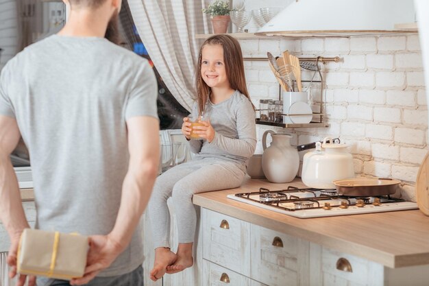 Happy birthday girl Loving father prepared surprise gift for his little daughter Smiling kid sitting in kitchen with glass of juice