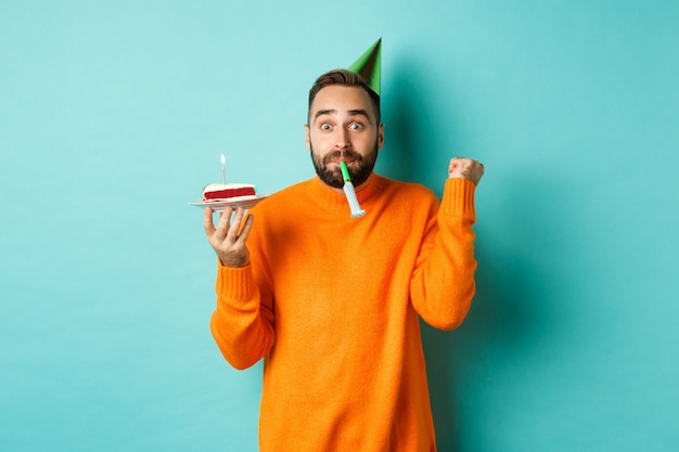 С днем рождения парень празднует, в шляпе для вечеринки, дует в свисток и держит торт ко дню рождения и делает