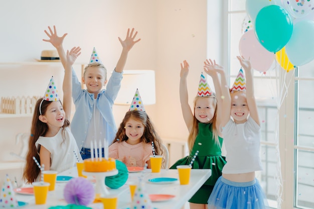 Happy birthday party Joyful small children have fun together raise arms and play with confetti pose at festive table wear party hats being on carnival enjoy holidays