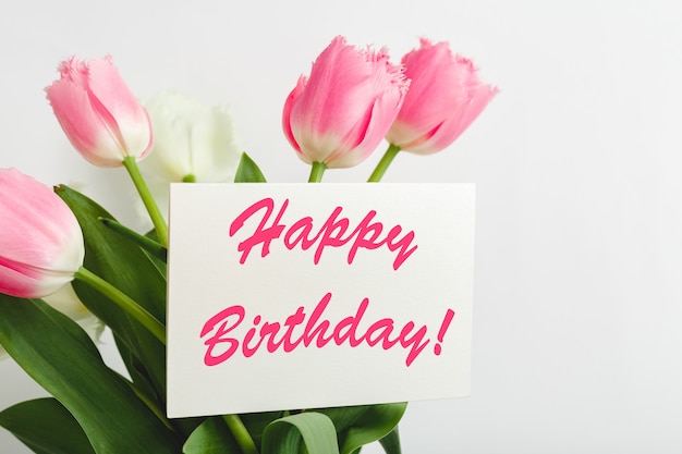 Happy Birthday text on gift card in flower bouquet. Beautiful bouquet of fresh flowers tulips with greeting card Happy Birthday on white wall