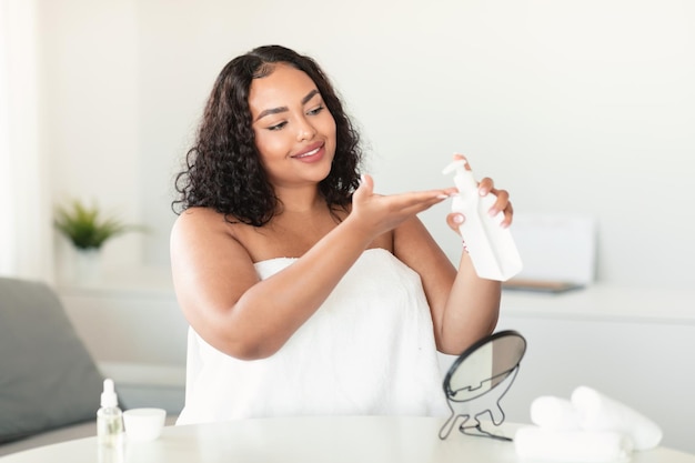 Photo happy black body positive lady applying moisturising lotion on hand holding bottle woman standing wrapped in towel