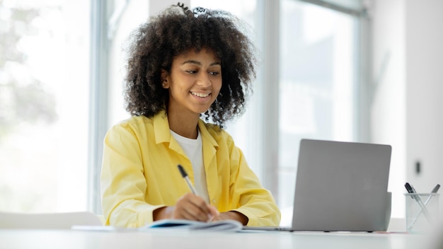 Happy black brunette woman using laptop smiling while working while sitting at workplace in modern o