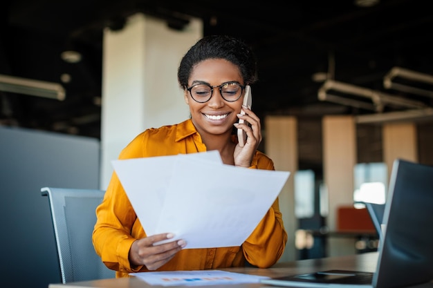 Happy black businesswoman talking on cellphone and checking documents in office working with papers at workplace