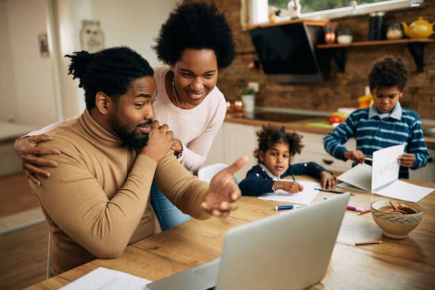 Happy black couple talking while man is working on laptop at home