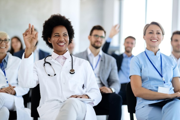 Happy black doctor raising her hand to ask a question during a\
seminar in convention center