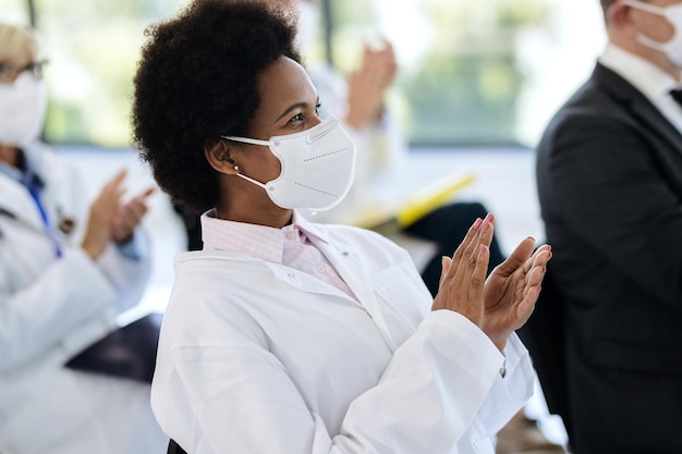 Happy Black doctor wearing face mask and applauding on a seminar in board room