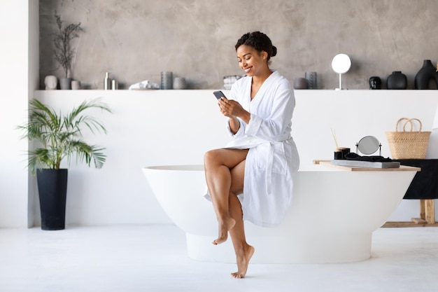 Happy black female using smartphone while sitting on bathtub in bathroom