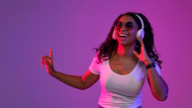 Happy black female wearing headphones and stylish sunglasses posing in neon light