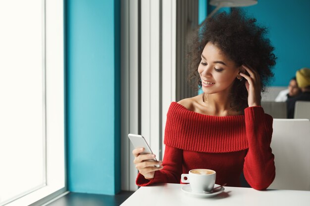 Happy black girl at cafe table typing on smartphone. Young woman enjoying hot coffee and surging the Web. Leisure, technology and urban lifestyle concept.