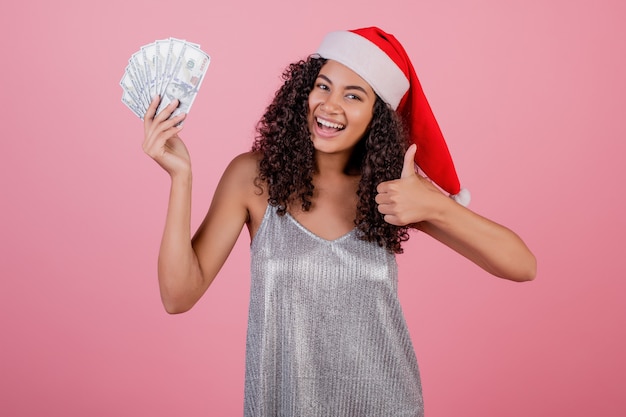 Happy black girl holding one hundred dollar bills wearing santa hat isolated 