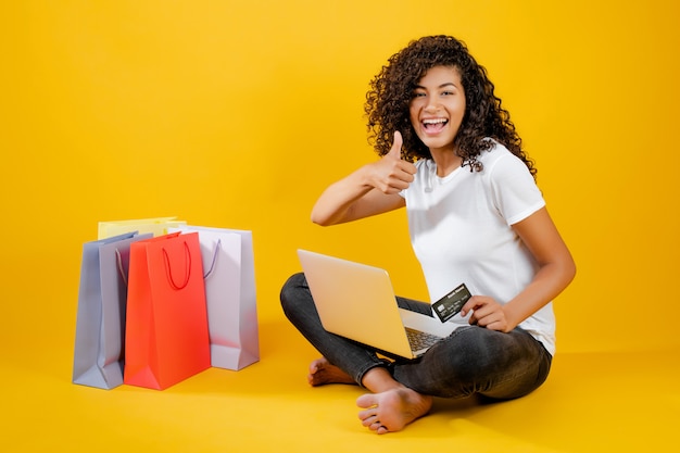 Happy black girl with colorful shopping bags sitting with laptop and credit card isolated over yellow