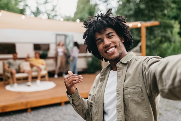 Happy black guy taking selfie near rv smiling at camera resting with his multiracial friends