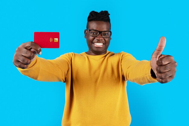 Happy black guy wearing eyeglasses holding credit card and showing thumb up