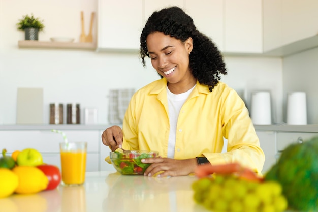 Happy black lady eating fresh vegetable salad enjoying healthy food and drink sitting in modern kitchen at home