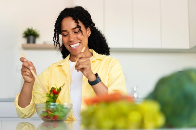 Happy black lady enjoying cooking preparing healthy vegetable\
salad and mixing ingredients in bowl standing in kitchen