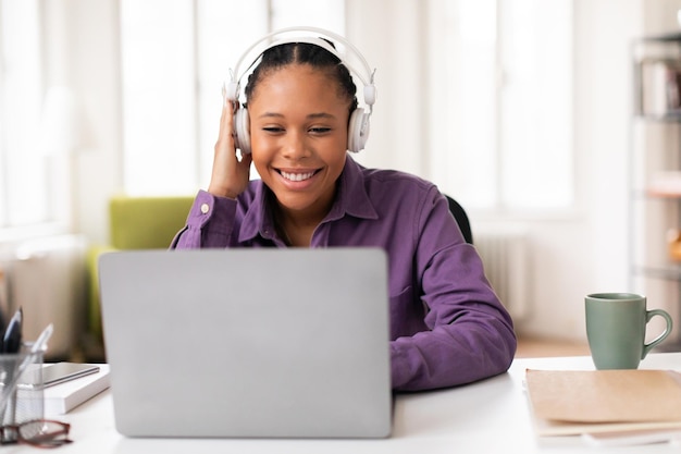 Happy black lady student looking at laptop and touching headphones