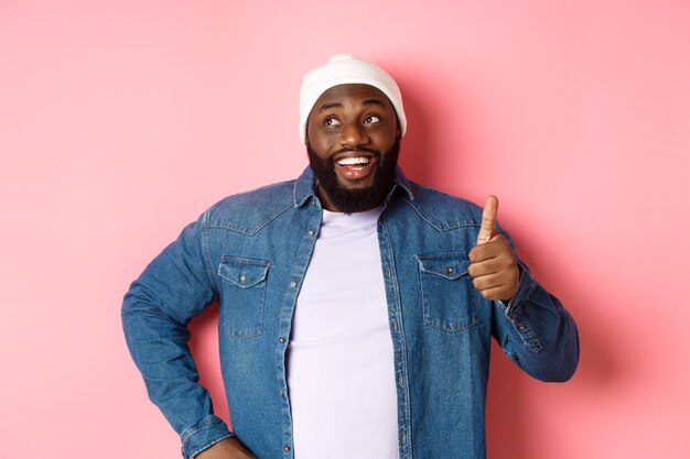 Happy Black man showing thumb-up, looking at upper left corner thoughtful, standing over pink background.