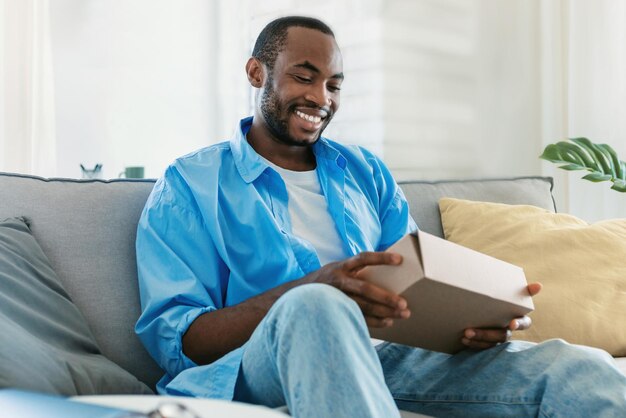 Happy black man unpacking delivery box sitting on sofa and smiling male customer opening package