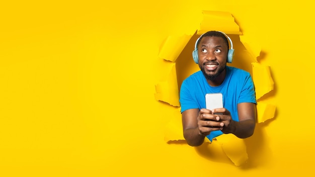 Счастливый темнокожий мужчина в наушниках и с помощью смартфона смотрит на свободное пространство через дыру в порванной желтой бумаге