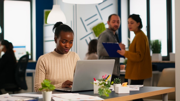 Happy black manager reading tasks on laptop and typing sitting at desk in busy start up office while diverse team analyzes statistics data in background. Multiethnic group talking about project