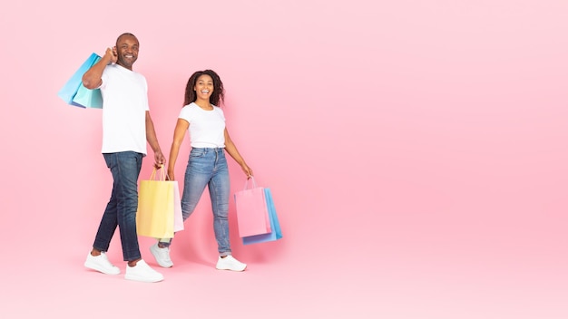 Happy black middle aged man and young woman walking with shopping bags over pink background copy space