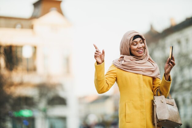 Happy black Muslim woman wearing a hijab and making video call with her smartphone while walking downtown.