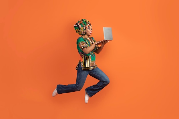 열린 노트북으로 점프하는 아프리카 의상을 입은 행복한 흑인 여성