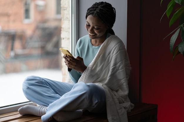 Happy black woman enjoying buying gifts ordering food and\
sitting by window with smartphone at home