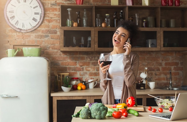 Happy black woman talking on phone and drinking wine while cooking healthy food in kitchen