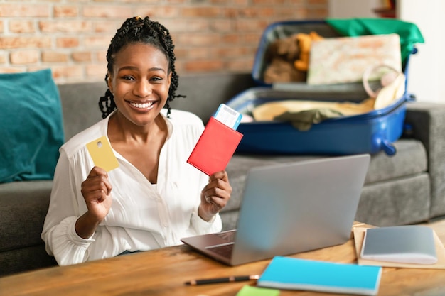 Happy black woman using laptop buying travel tickets holding passport and credit card sitting with