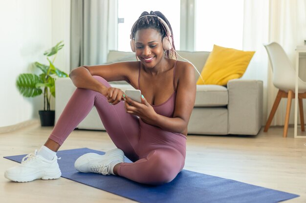 Photo happy black woman using smartphone and exercising wearing headphones sitting on fitness mat during online workout