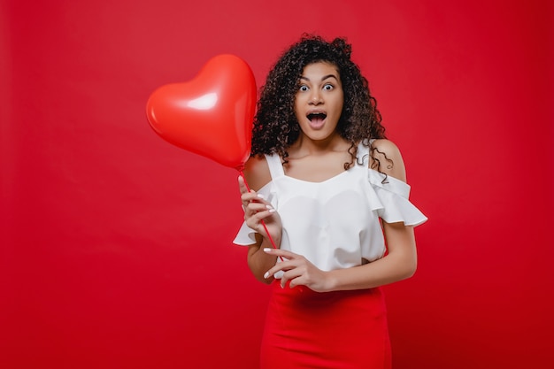 Happy black woman with heart shaped balloon wearing skirt isolated on red wall