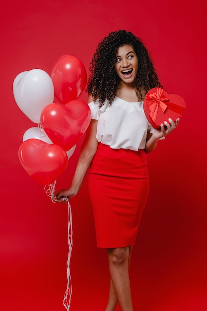 Happy black woman with heart shaped gift box and colorful balloons isolated on red wall