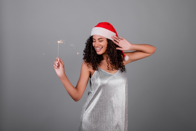 Donna di colore felice con le stelle filante scintillanti che portano il cappello e il vestito di natale isolati sopra grey