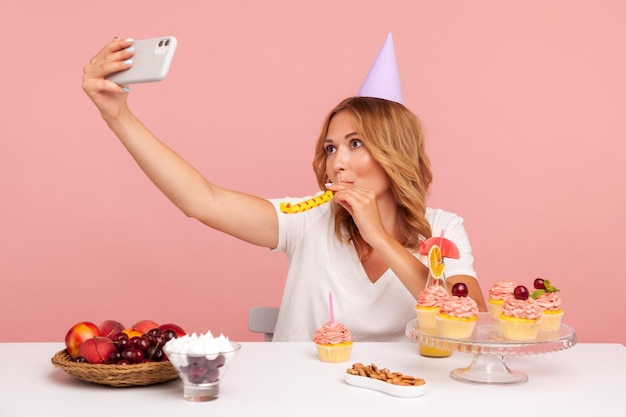 Happy blogger broadcasting livestream with phone and blowing horn to camera wearing white Tshirt and party cone celebrating birthday online Indoor studio shot isolated on pink background