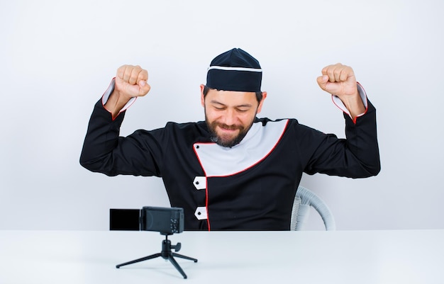 Happy blogger chef is raising up his fists by sitting in front of his mini camera on white background