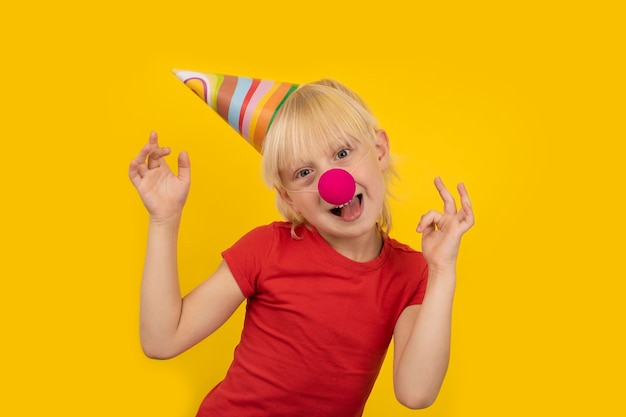 Happy blond boy celebrating his birthday Child in party hat red clown nose on yellow background