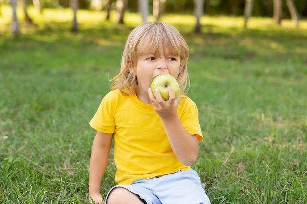 a happy blond boy is sitting on the lawn in a yellow T-shirt biting a green apple