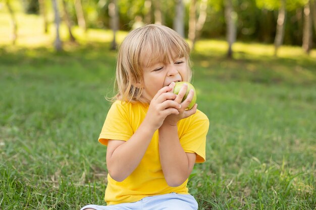 a happy blond boy is sitting on the lawn in a yellow T-shirt biting a green apple