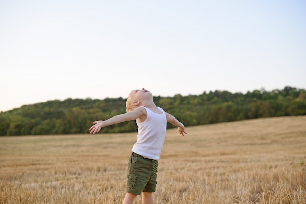 Happy blond boy stands with his arms apart and head up in a mowed wheat field