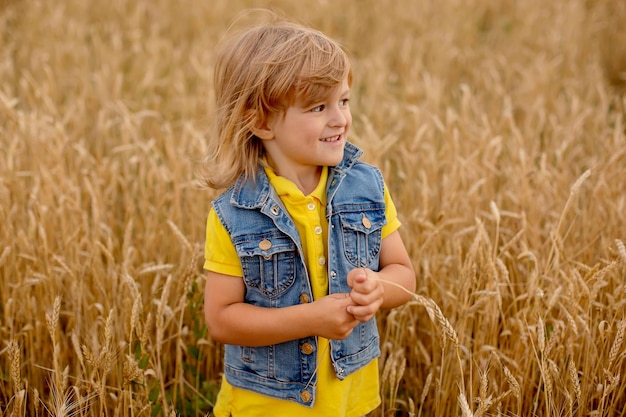 a happy blond boy in a yellow jacket and a denim vest is standing in a wheat field