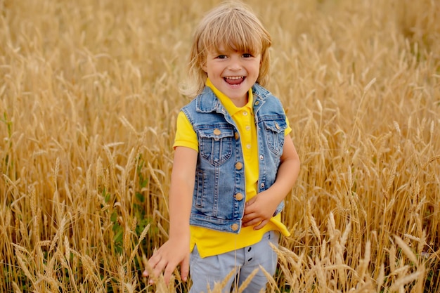 a happy blond boy in a yellow jacket and a denim vest is standing in a wheat field