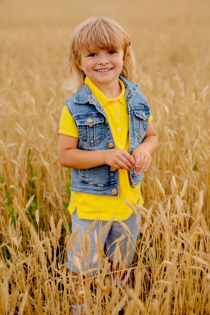 a happy blond boy in a yellow jacket and a denim vest is standing in a wheat field