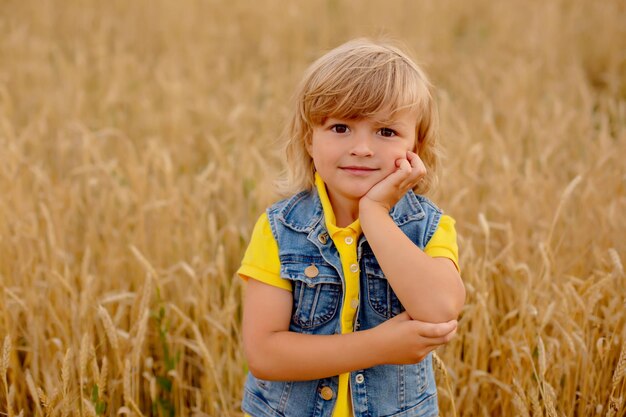 a happy blond boy in a yellow jacket and a denim vest is standing in a wheat field