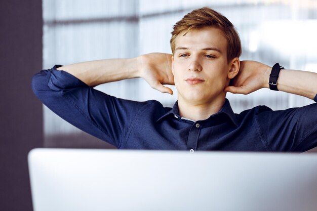 Happy blond businessman and programmer stretching hands after working hard with computer. Startup business leads to success