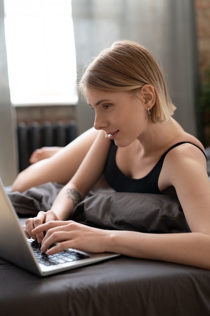 Happy blond girl in leotard surfing in the net while lying on bed after sleep and scrolling through online videos or posting her new photos